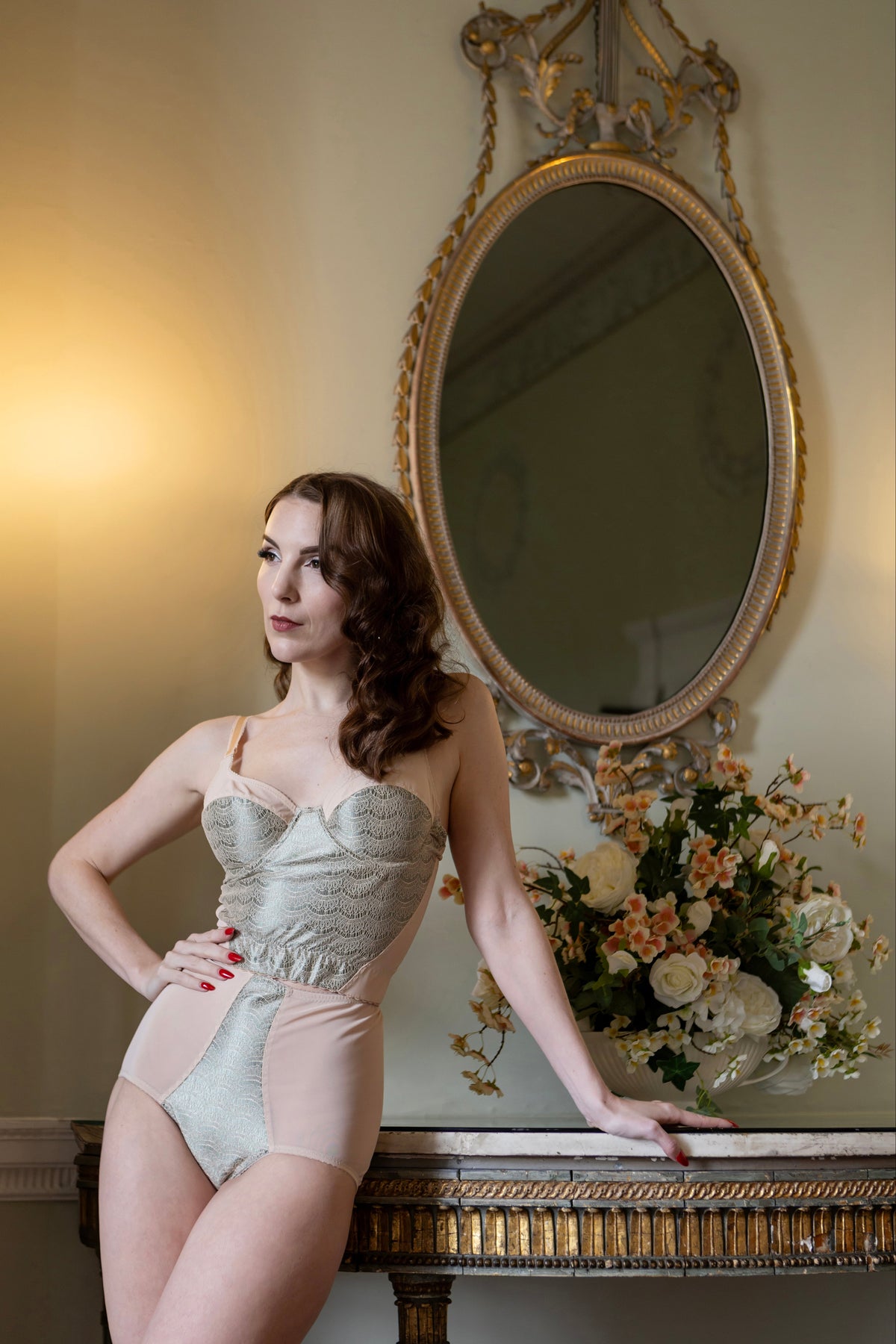 A woman models a light beige and peach-colored lace and mesh bodysuit while leaning on a decorative console table in front of an ornate gold mirror.