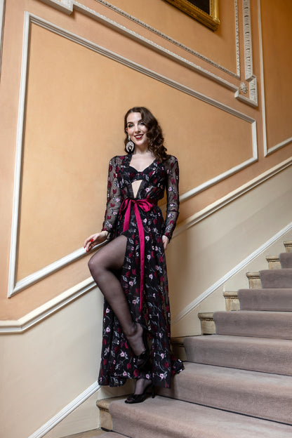 A woman posing on an indoor staircase wearing a long-sleeved black floral maxi dress with sheer mesh sleeves, a keyhole cutout at the chest, and a prominent magenta pink waist tie.
