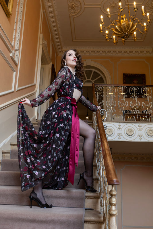 A woman in a vintage-style black floral dress with a bright pink sash and sheer sleeves stands on an indoor staircase, holding out the skirt to show the flow. The setting features ornate cream walls, a large chandelier, and a wooden handrail.