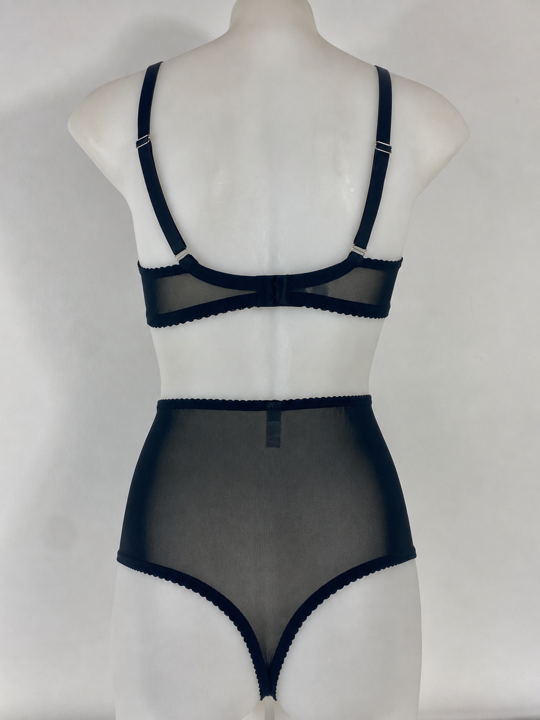 A two-piece lingerie set consisting of a sheer black mesh bralette with solid black trim and adjustable shoulder straps, paired with a matching high-waisted sheer mesh brief, displayed on a white mannequin torso against a light gray background.